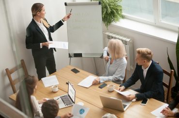 Businesswoman ceo boss in suit presenting corporate strategy pointing on flip chart at group meeting, business coach leader giving presentation explaining team goals in conference room at training